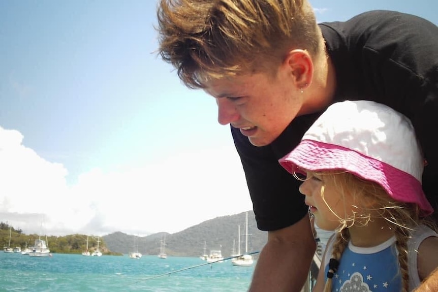 a young man in a black t shirt bends down to a young girl standing against a beautiful blue ocean