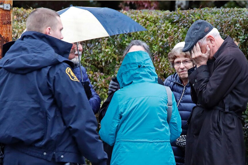 Man stands with head in his hands and people gather on a street corner near a synagogue where a shooter killed many.