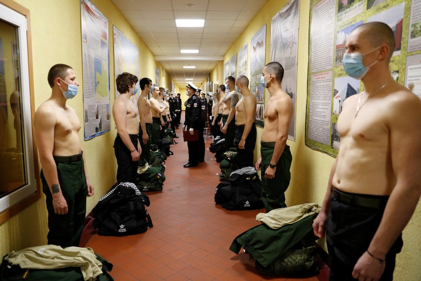 A group of man without their shirts on stand against a wall as they are inspected by a man.