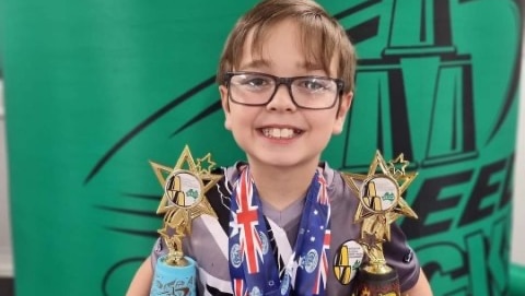 boy with spectacles, smiling, holding trophies in front of green banner in background