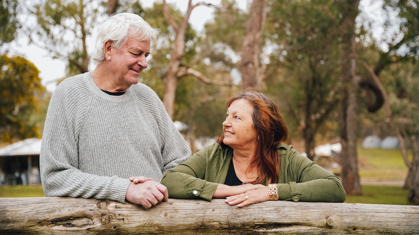 A man and woman lean against a wooden fence post smiling.