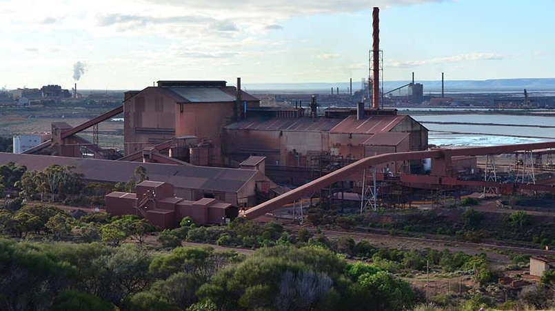 The rusty red industrial building with conveyers and chimneys, and a rail track and puffs of steam from a chimney.