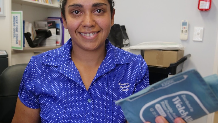 Dani Beezley, smiles at the camera, wearing blue shirt with white dots, holding a blood pressure cuff, bookshelf, office behind.