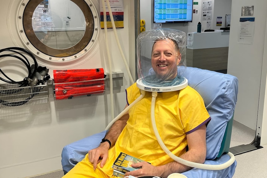 A man in a yellow hospital gown sits in a chair smiling at the camera. He is wearing hyperbaric chamber headwear