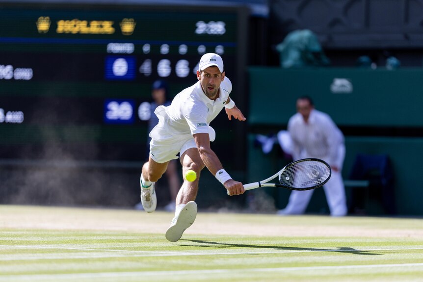 Novak Djokovic lunges forward to hit a low backhand return at Wimbledon as dust rises from the grasscourt.
