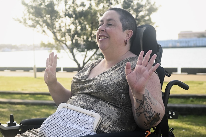 A woman in a wheelchair, smiles and holds up her hands during a conversation.