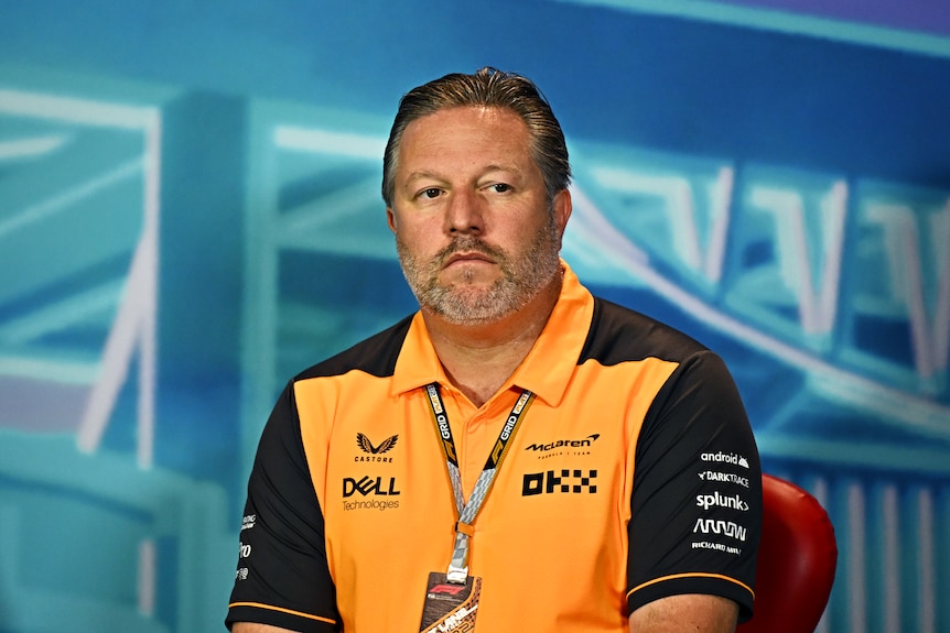 Man in an orange shirt sitting at a press conference frowning.