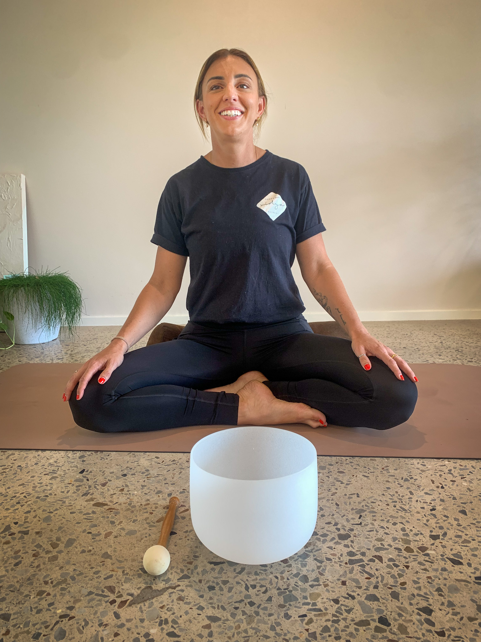A blond woman sits on a yoga mat in a yoga pose. She has a drum in front of her
