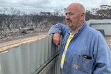 A bald man wearing a denim tracksuit looking over his fence towards burnt spindly trees