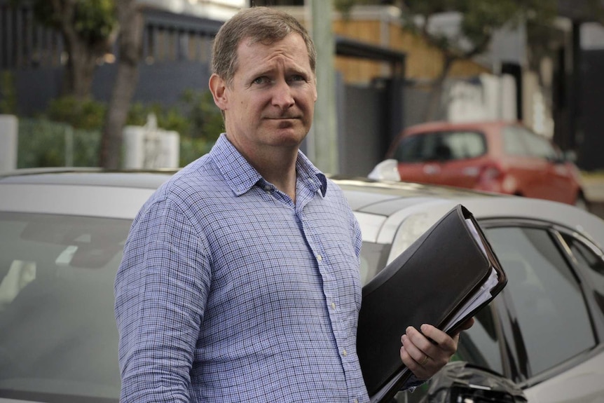 Lenny Willis standing on the street in front of a parked car and holding a document wallet