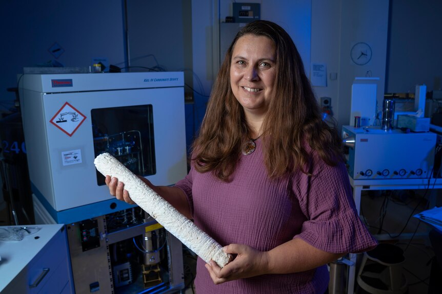 A woman in a blue lit lab, holds a large white coral core, she has long hair and wears purple. 