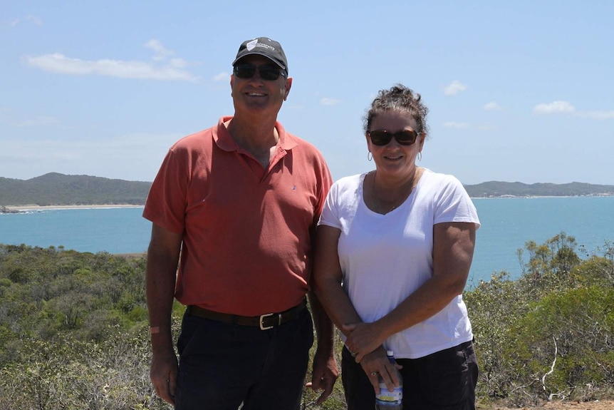 A man and woman stand at a lookout over the ocean and smile at the camera.