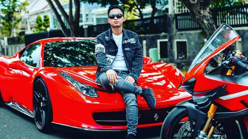 A man wearing a leather jacket poses on top of a red luxury vehicle 