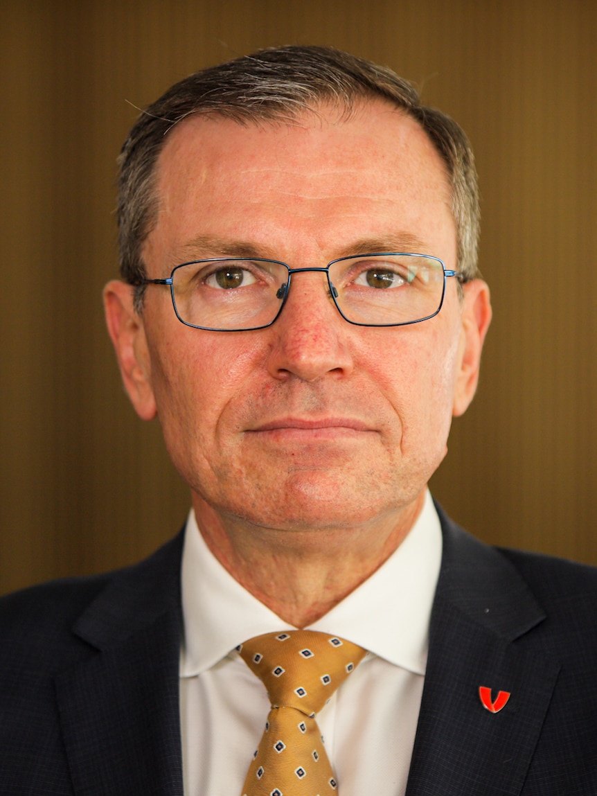 Middle aged man with short hair and glasses looks directly at the camera, brown background 