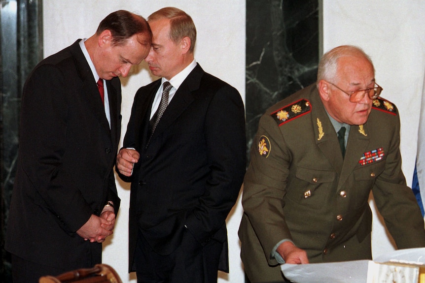 A man bows his head to listen to Vladimir Putin, while another man in military uniform shuffles some papers