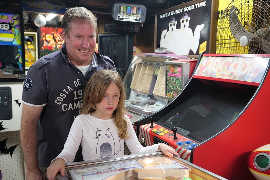 Lloyd coaches his daughter Samantha behind a pinball machine in his crowded basement.