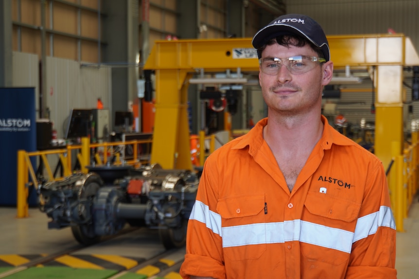 A man wearing an orange hi-vis long sleeve shirt, safety glasses and a hat standing in front of train parts.