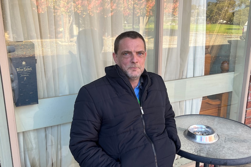 Man sits in front of motel room with puffer jacket on and hands in pockets. 