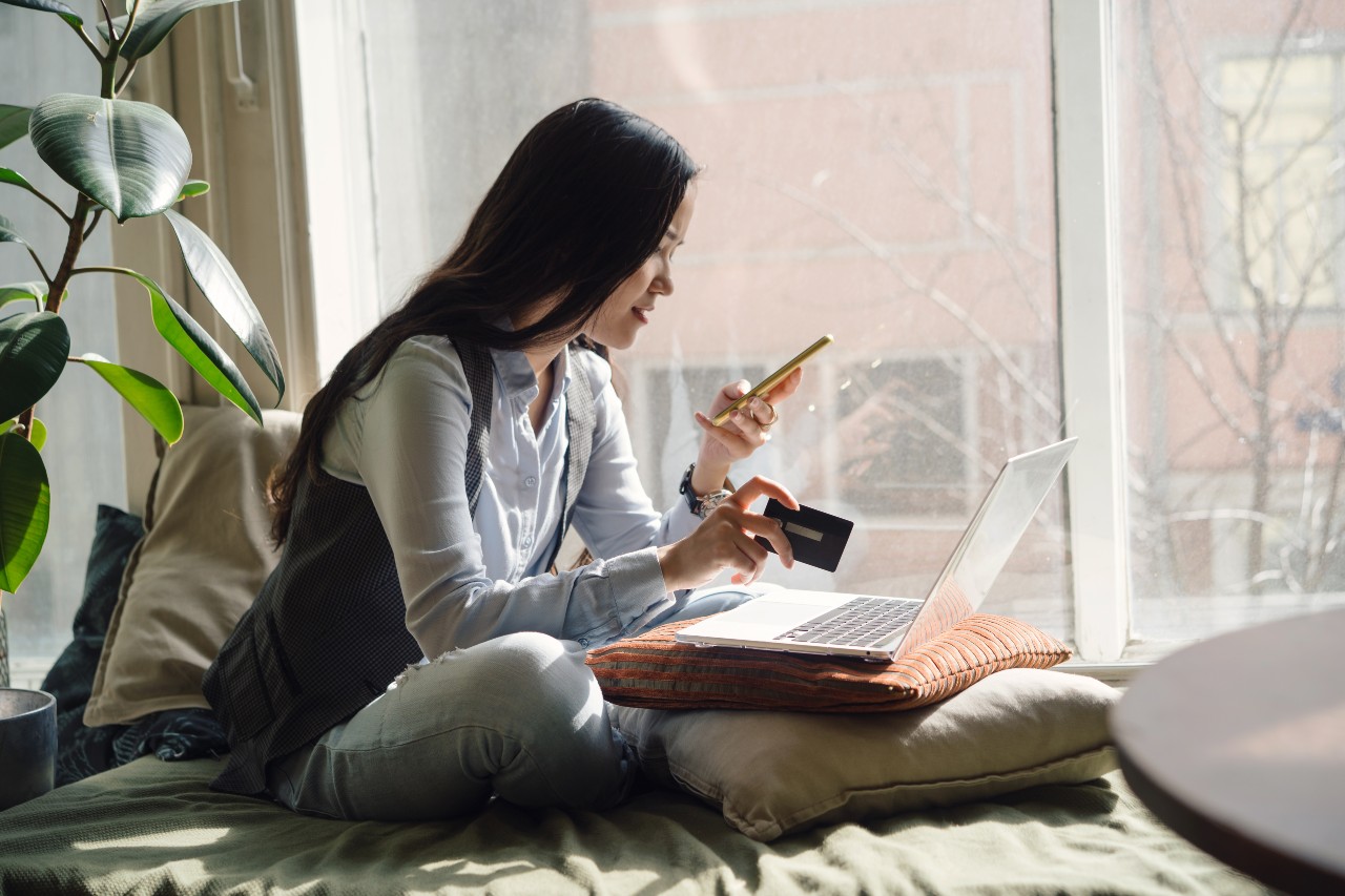 A woman sits cross-legged on a couch with her laptop in front of her, holding up her phone and credit card.