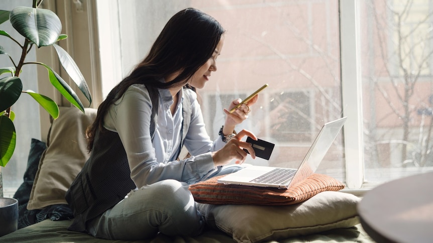 A woman sits cross-legged on a couch with her laptop in front of her, holding up her phone and credit card.