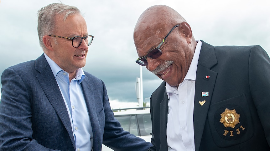 Two men in suits shake hands as they meet ouside under a grey sky.