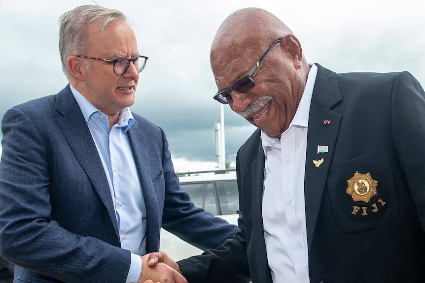 Two men in suits shake hands as they meet ouside under a grey sky.