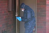 A mid-shot of a police forensics officers at the front door of a red brick house wearing a blue jumpsuit, gloves and face mask.