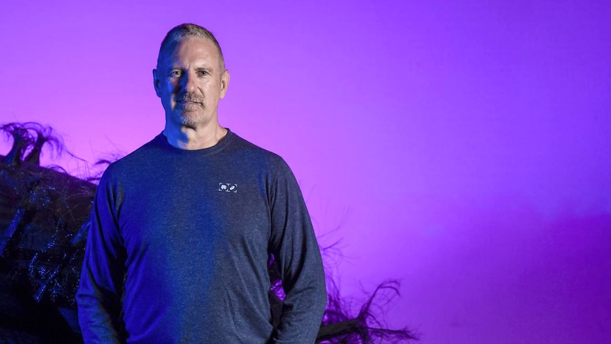 A man stands on a stage with a purple backdrop and some kind of large black hairy thing behind him