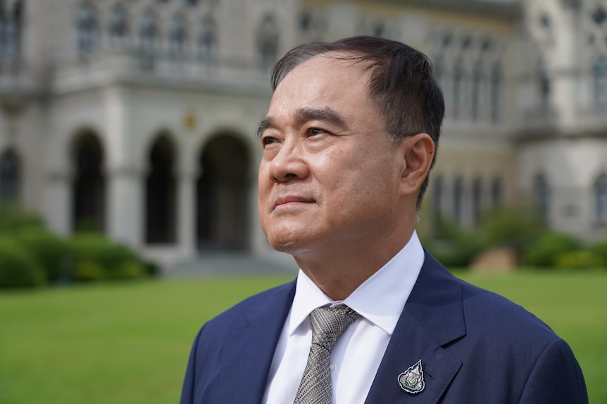 A man wearing a suit and tie looks upwards, standing on grass in front of an ornate old building