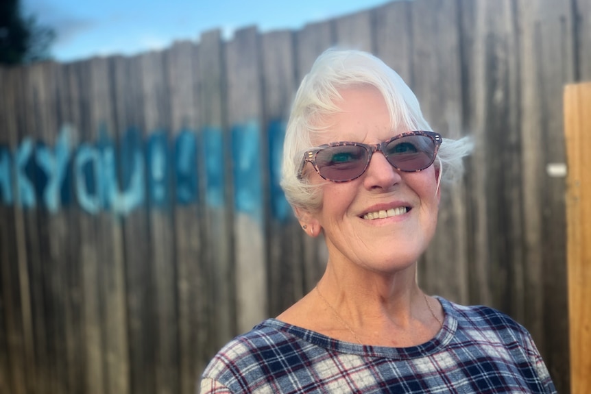 A woman wearing sunglasses smiles in front of a fence with blue graffiti on it.