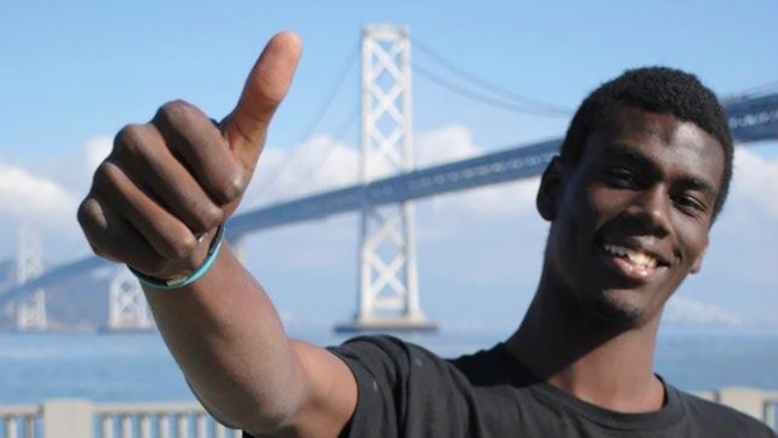 A man stands smiling in front of a bridge holding up a thumbs up. 