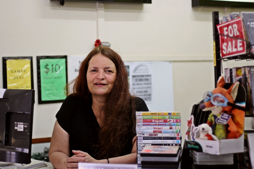 A woman sits at the counter of a video store, surrounded by stacks of DVDs