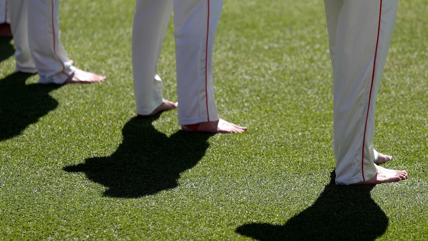 The barefoot feet of three cricketers as they stand on grass.