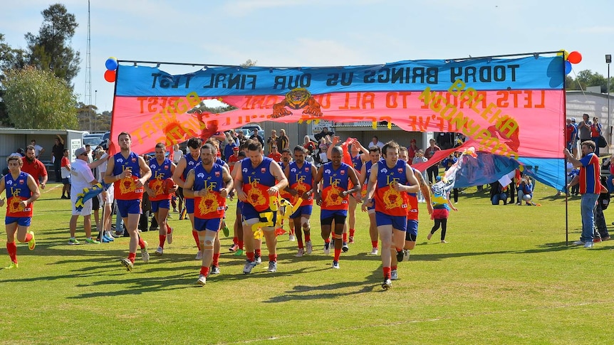 Football players wearing blue and red run through a blue and red banner at a country football oval.