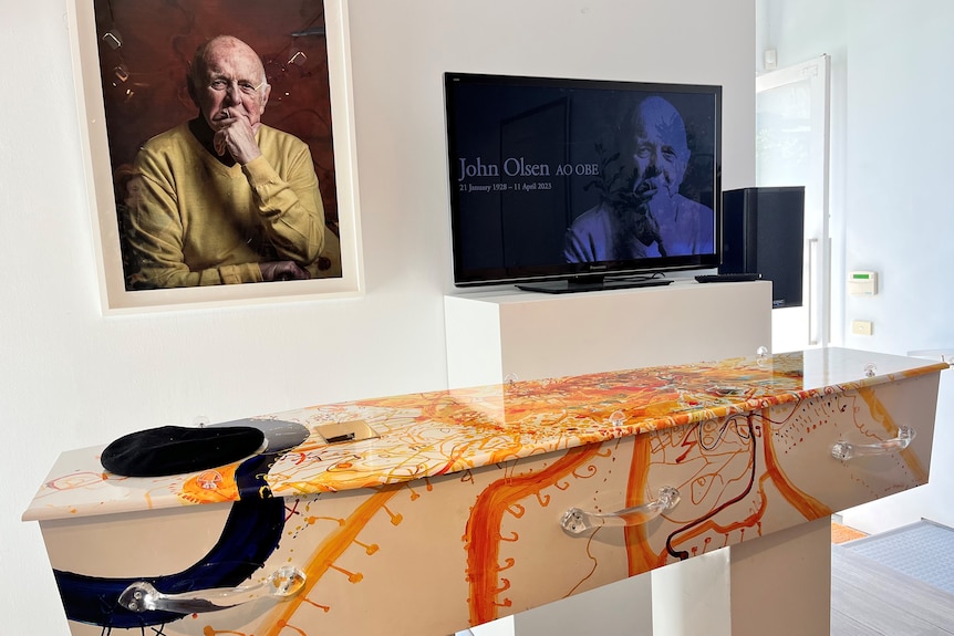 A coffin decorated with golden colours sits below a painting of an old man in a pondering pose