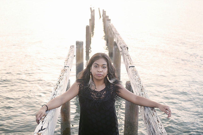 A woman stands with arms resting on an old wooden jetty over water.