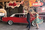 Mick twirls Di around as they dance, her 1950s skirt puffing out around her, in front of a red ford
