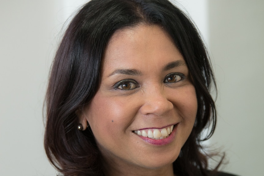 A woman with shoulder-length brown hair, a black suit jacket and purple floral top smiles widely.