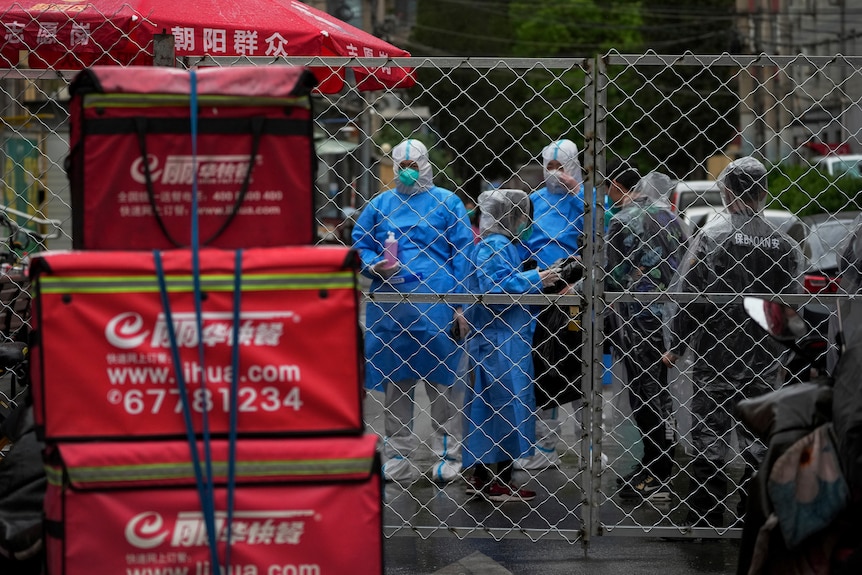 Health workers wearing protective gear chat with security guards behind a barricaded fence