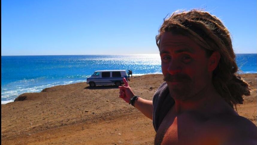 Adam Coleman takes selfie while pointing at their van on a beach.