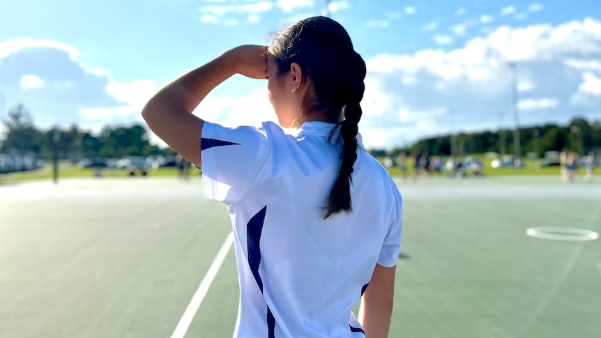 A young girl with dark hair tied back, looks over a netball court. The photo is taken from behind her.