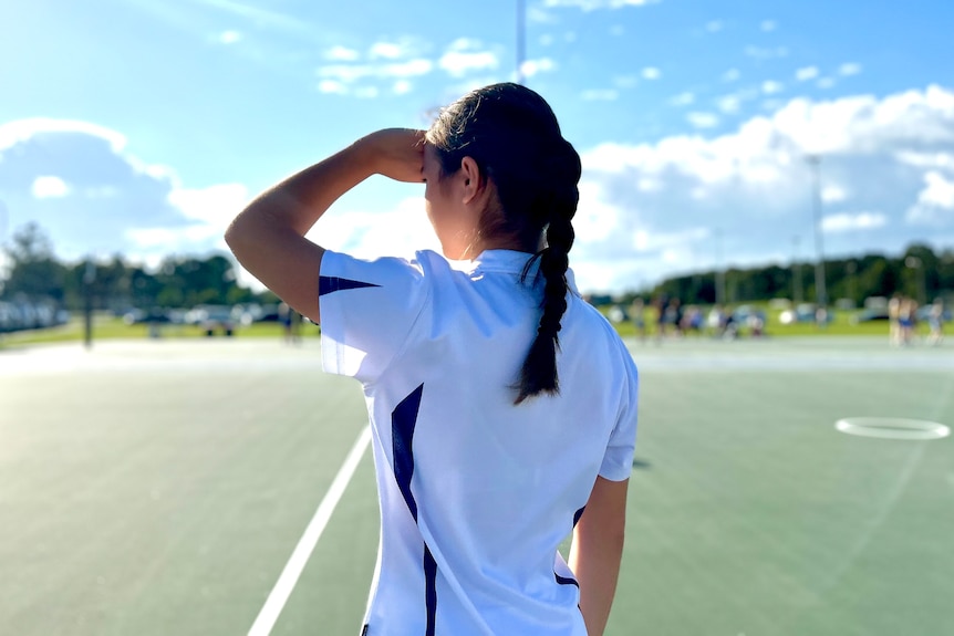 A young girl with dark hair tied back, looks over a netball court. The photo is taken from behind her.