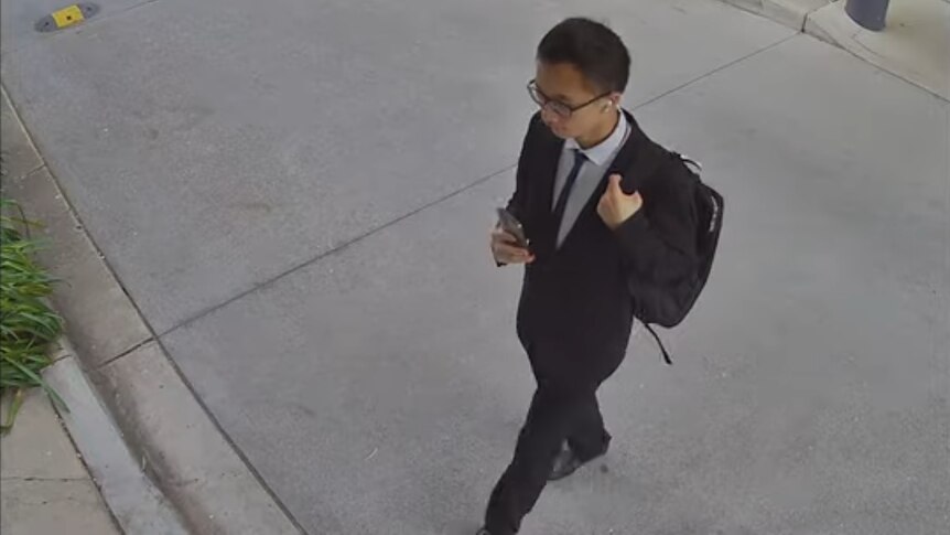 A man in a suit holds a phone up, captured from above on CCTV.