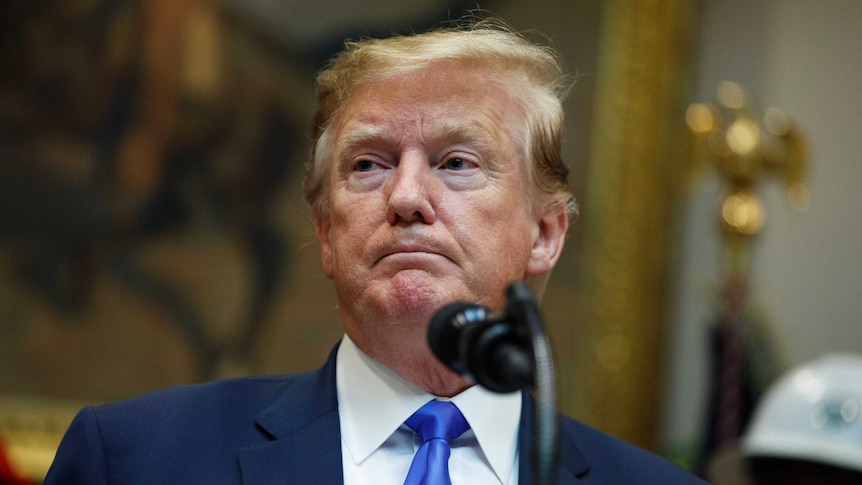 Donald Trump wears a navy suit and looks glum while standing in front of a microphone