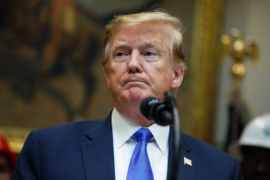 Donald Trump wears a navy suit and looks glum while standing in front of a microphone