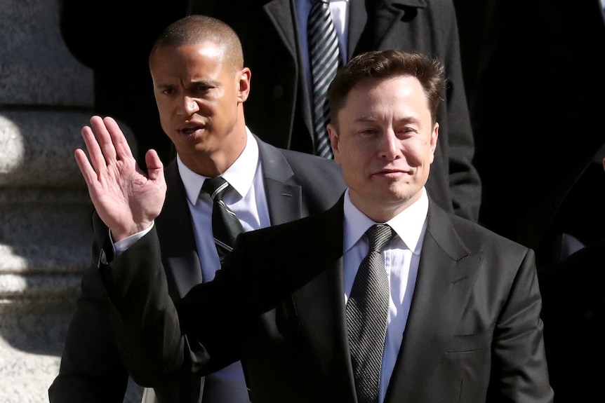 Wearing a black suit, Elon Musk waves as he walks on the steps of the courthouse.