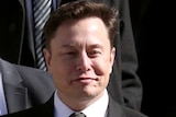 Wearing a black suit, Elon Musk waves as he walks on the steps of the courthouse.