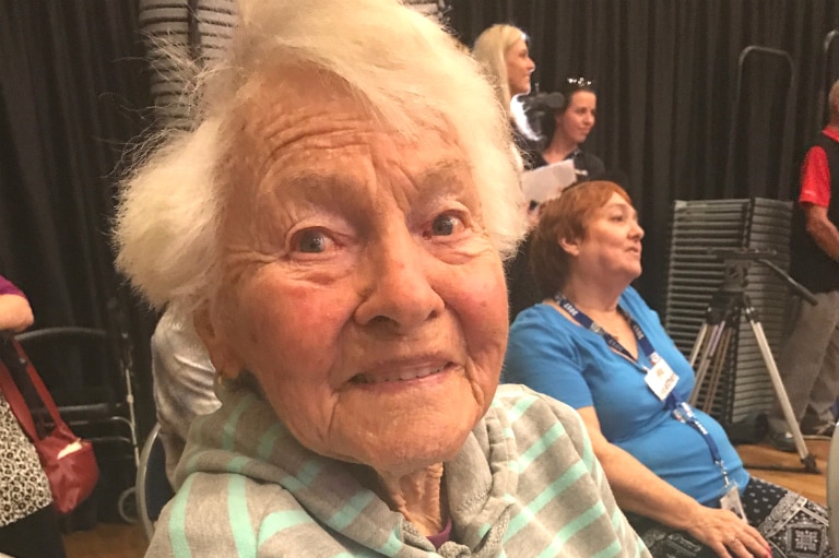 A 93 year old woman sits in a wheelchair looking at the camera.