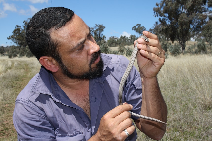 A man with a short beard holds a thin snake while crouched in grass.