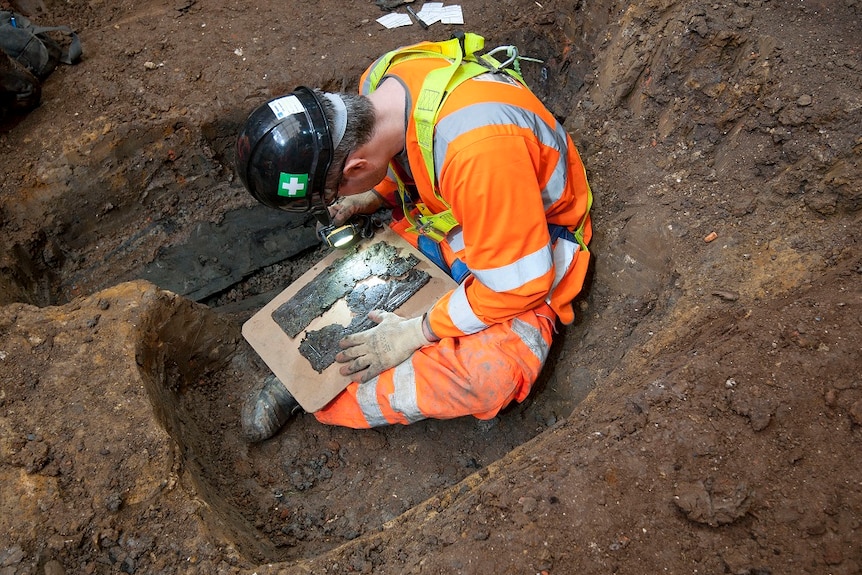 A man in high visibility safety gear sigs in a clay pit, examining the pieces of a coffin plate with a torch.